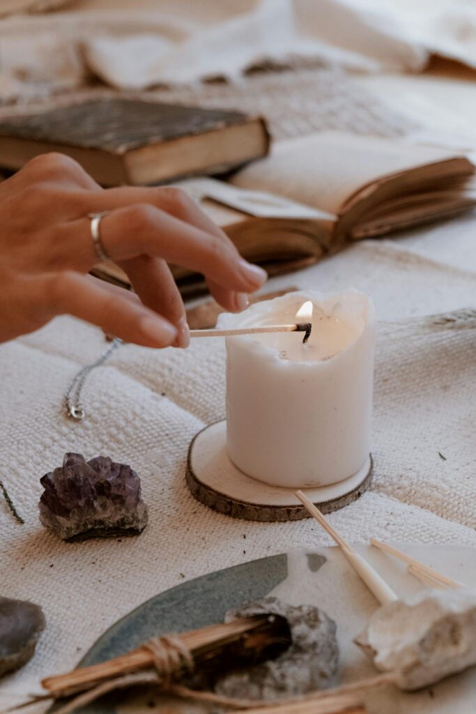 A hand lighting a candle with a matchstick, surrounded by books and crystals on a cozy setup.